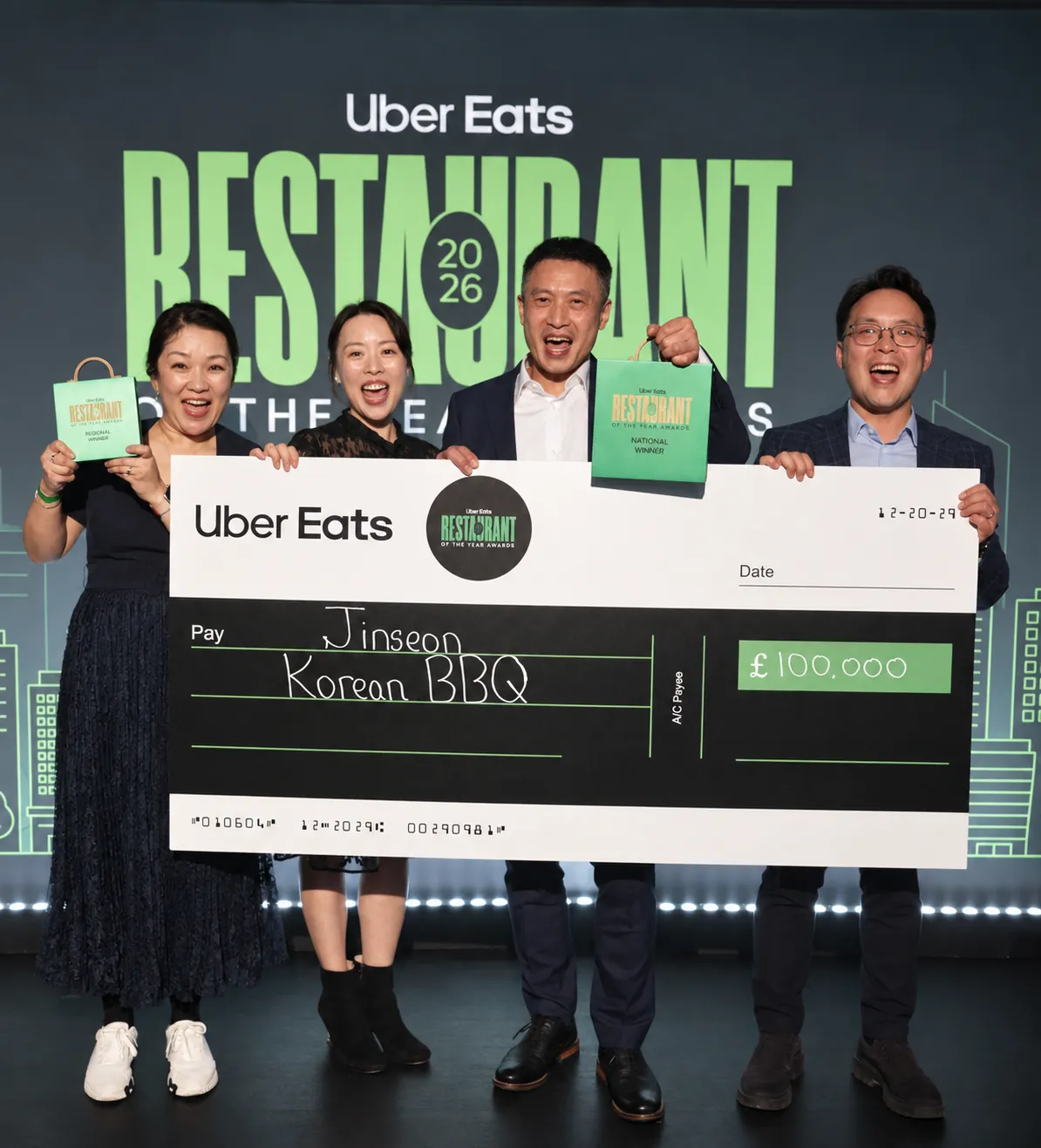 Four of the Jinseon family on stage at the awards night, cheering and holding up a giant cheque made out to Jinseon Korean BBQ, with the Regional Winner and National Winner awards in hand.