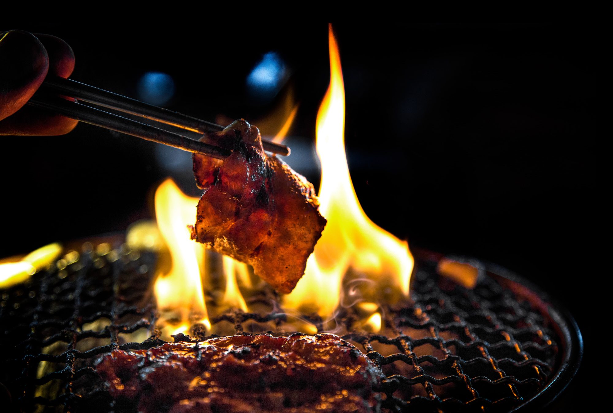 Chopsticks lifting a piece of marinated meat above open flame on the charcoal grill