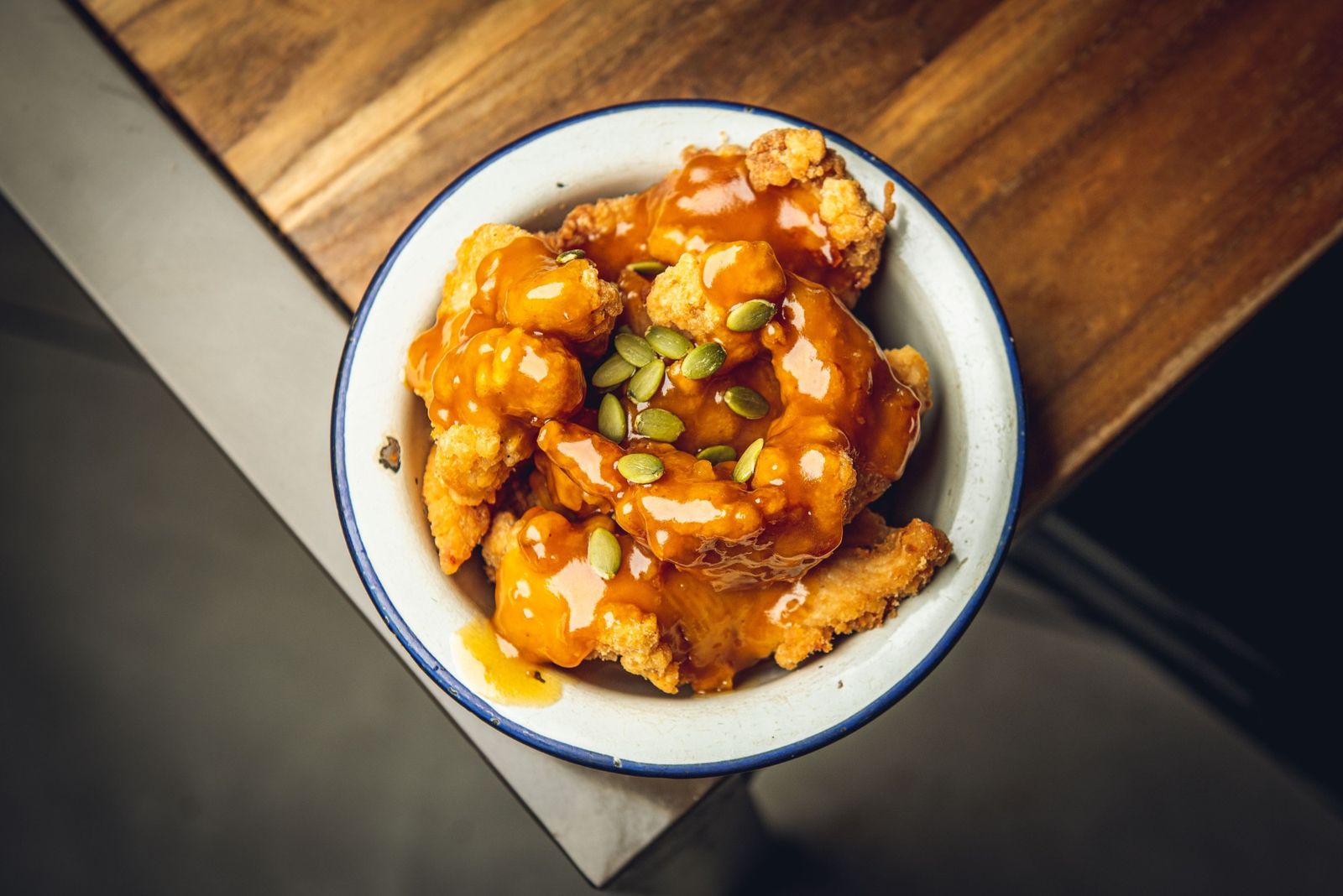 Korean fried chicken in a glossy sweet glaze, scattered with pumpkin seeds, in an enamel bowl