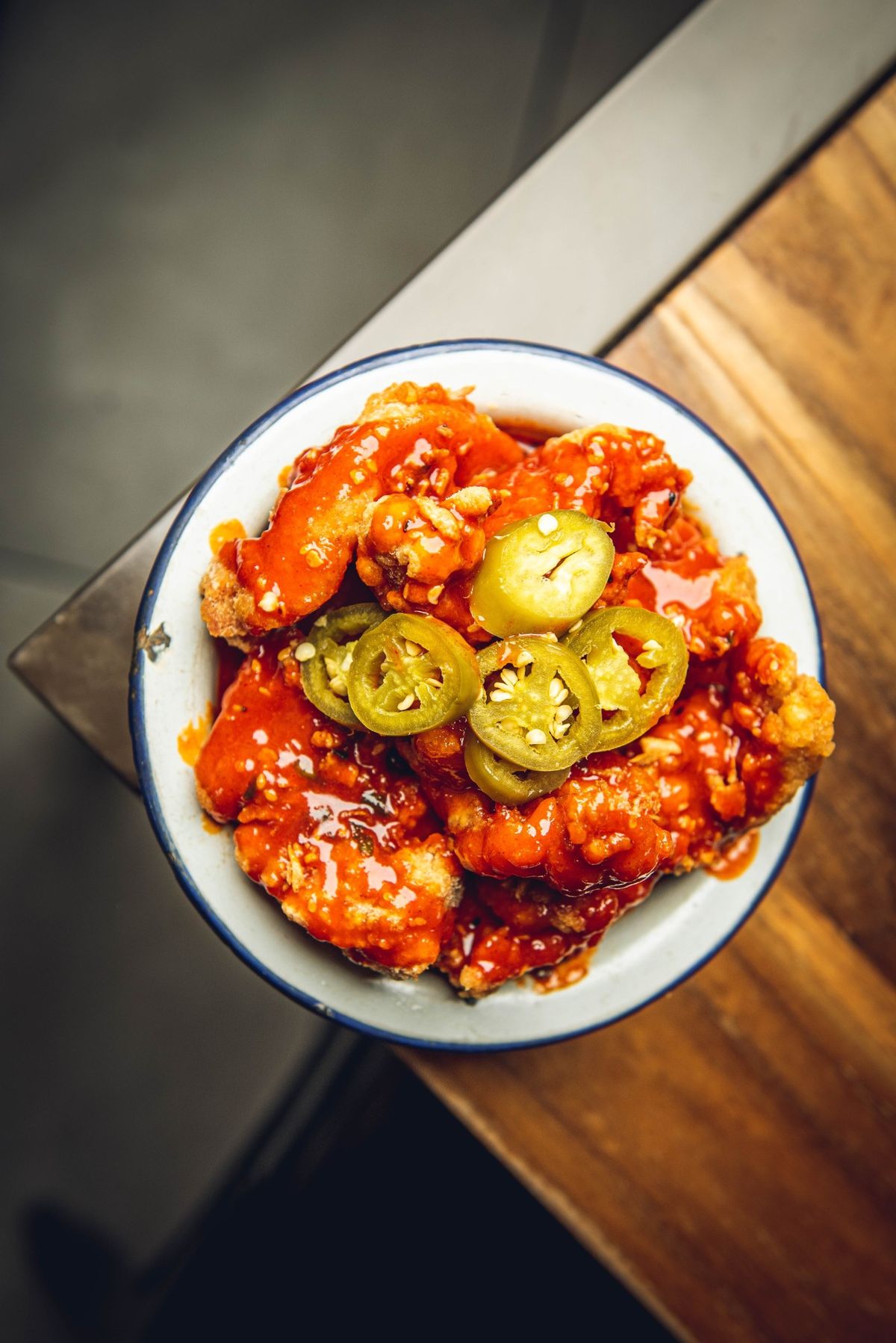 Korean fried chicken in a glossy red chilli glaze, topped with sliced jalapeños, in an enamel bowl