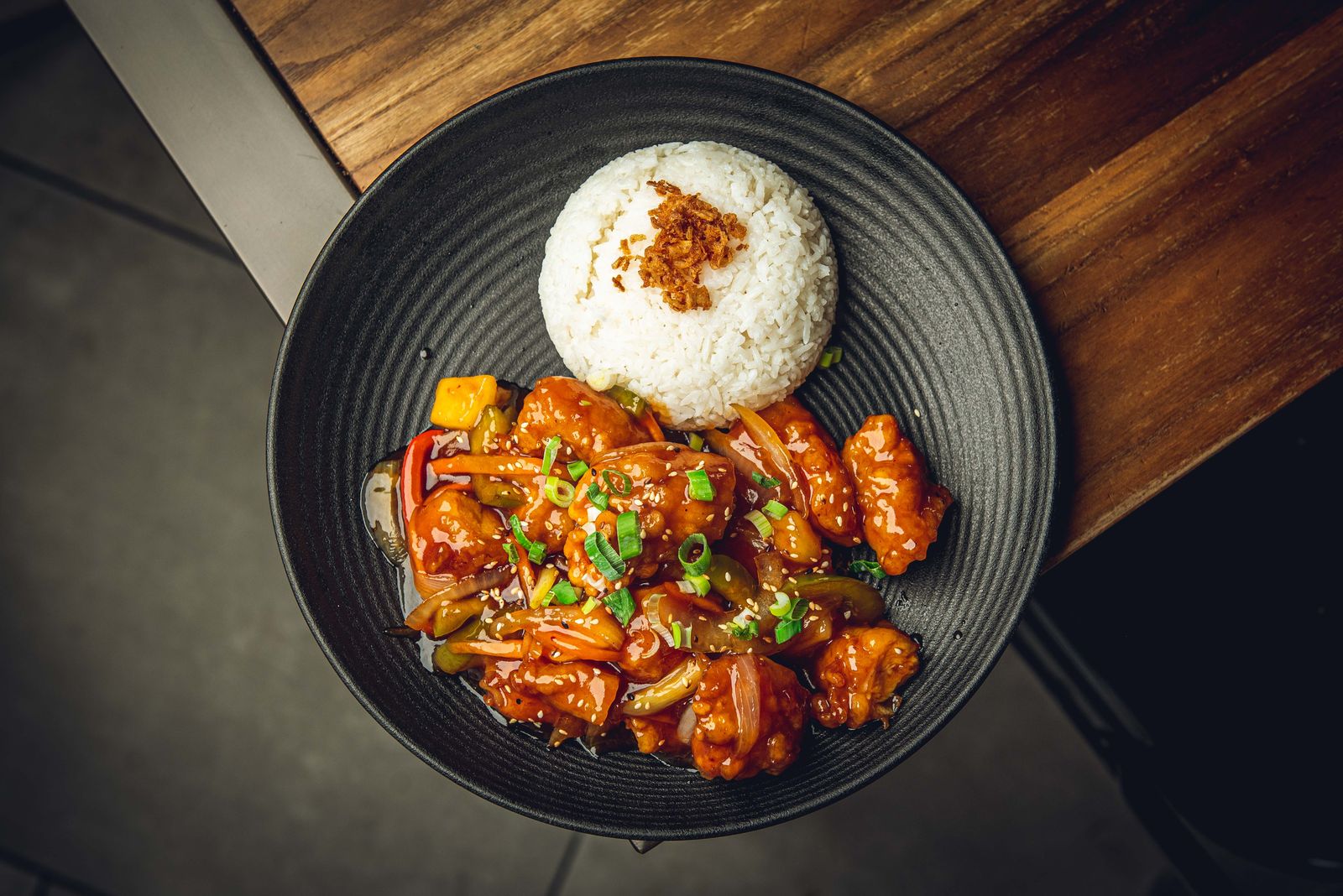 Korean sweet and sour crispy chicken with peppers, onion and pineapple, beside a dome of rice in a black bowl