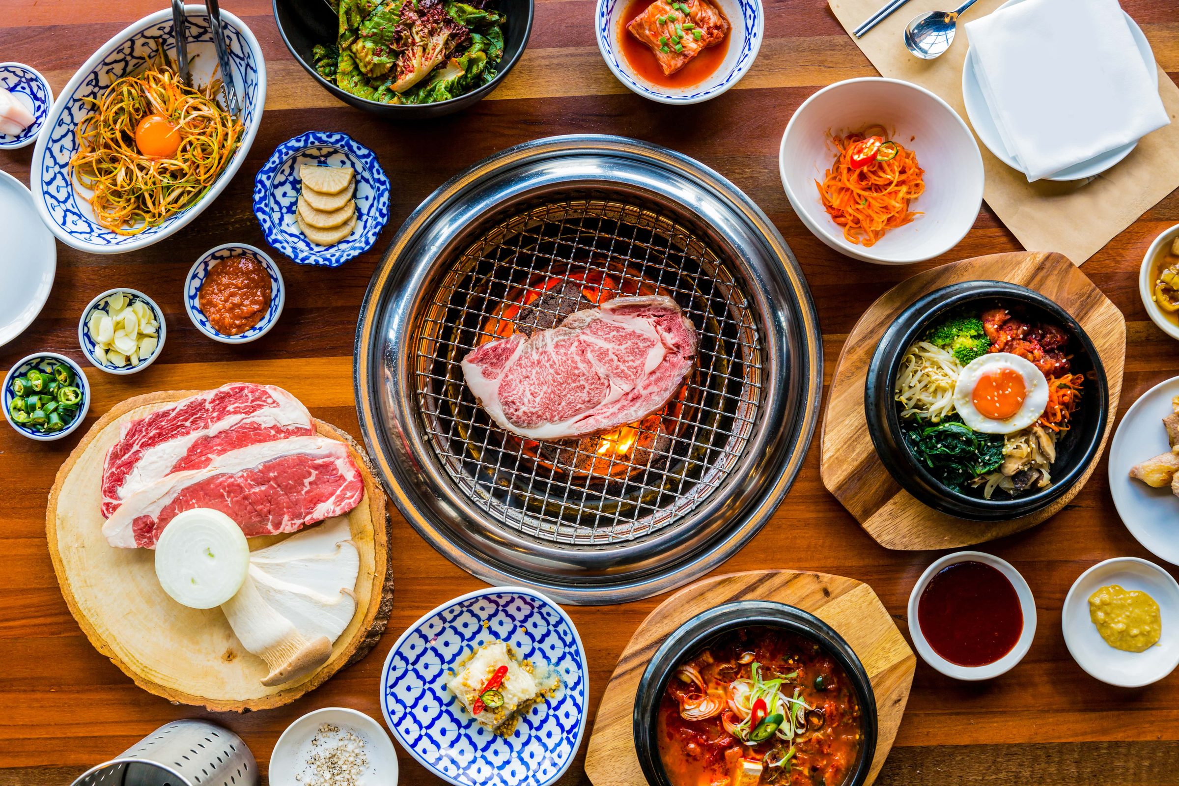 Looking down on a Jinseon table: the charcoal grill set into the wood with marbled beef searing on the grate, ringed by banchan, ssam lettuce, bibimbap and dipping sauces