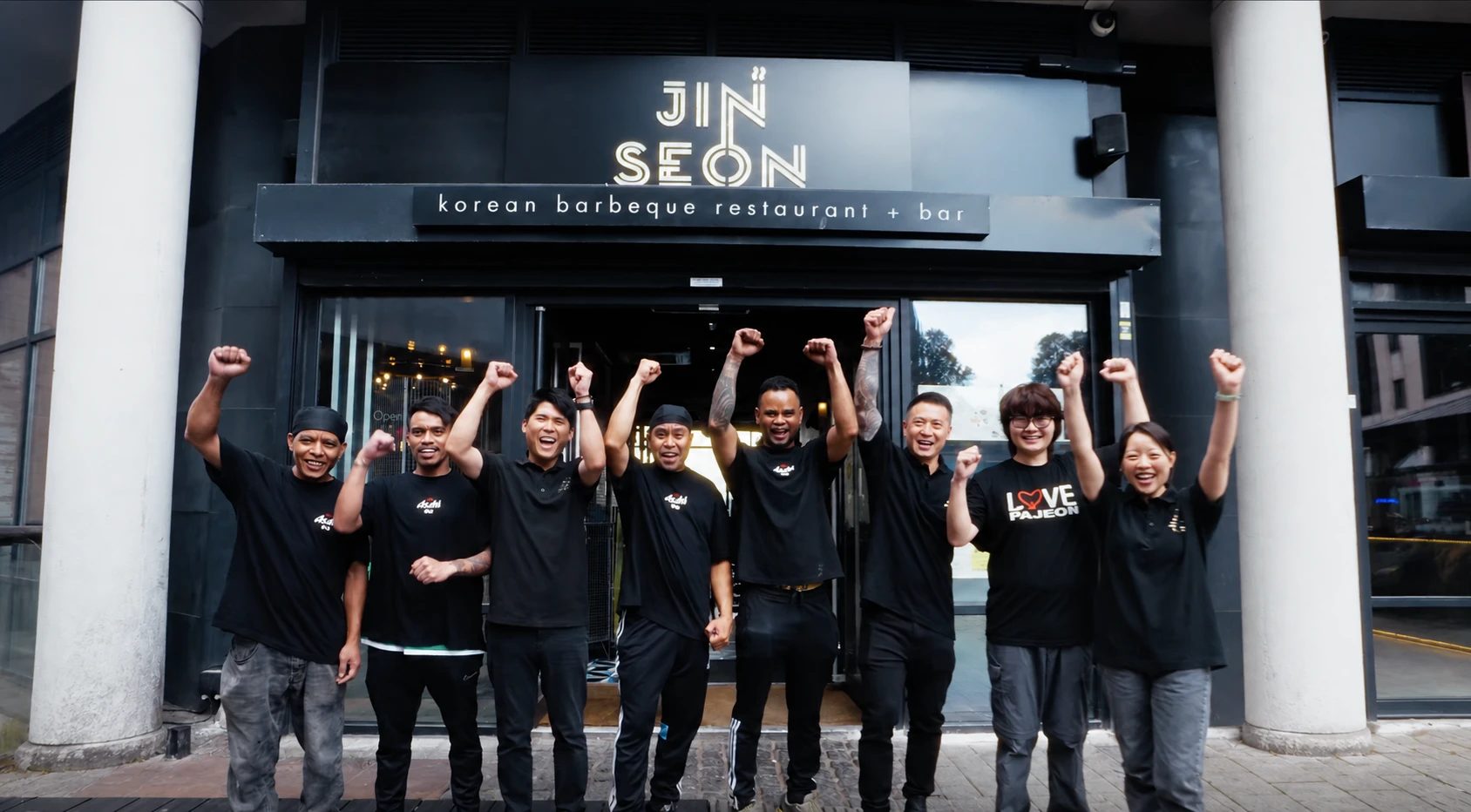 The Jinseon team outside the restaurant under the Jinseon Korean barbeque restaurant + bar sign, cheering with their fists in the air.